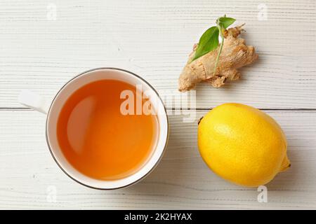 Tabletop view, porcelain cup of freshly brewed tea, dry ginger root with green sprout and whole lemon next to it, placed on white boards desk. Stock Photo