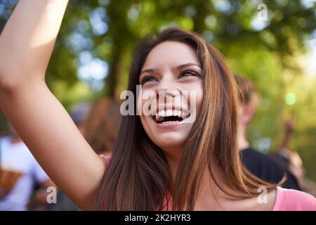 She's loves this song. A beautiful young woman smiling and enjoying music at a festival with arm raised in the air and crowd in the background. Stock Photo