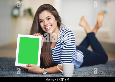 The best site youll ever find. Portrait of a smiling young woman lying on the floor at home holding up a digital tablet with a chroma key screen. Stock Photo