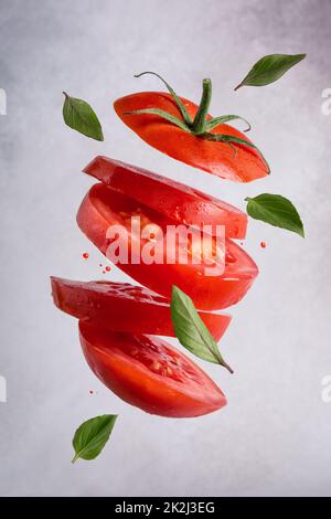Healthy and tasty vegetarian food. Flying ripe cut juicy tomato with fresh basil leaves on grey concrete background Stock Photo