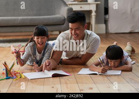 Being a father. Shot of a cheerful father and his two children doing homework together while lying on the floor at home during the day. Stock Photo
