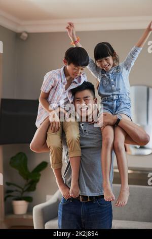 You guys have gotten heavier. Portrait of a cheerful young father holding his two children on each shoulder at home during the day. Stock Photo