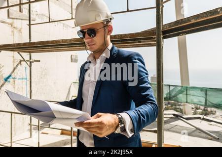 Man architect wearing formal suit and hard hat during building construction control holding a blueprints on a construction site Stock Photo