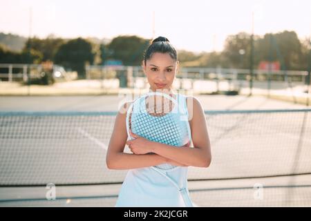 I spend most of my time on the court. Shot of an attractive young woman standing alone outside and posing with a tennis racket. Stock Photo