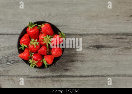 Tabletop view - small black ceramic bowl with freshly picked strawberries, on gray wood desk. Space for text on the right. Stock Photo