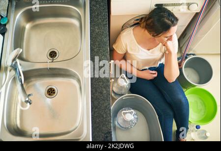 Worried about the water. High angle shot of a beautiful young woman feeling depressed about the water crisis whilst sitting on the floor in the kitchen at home. Stock Photo