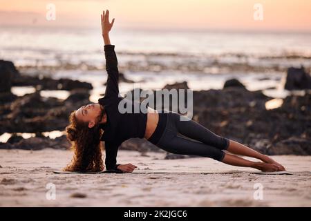 Breathe through each pose. Shot of an attractive young woman doing yoga alone on the beach at sunset. Stock Photo