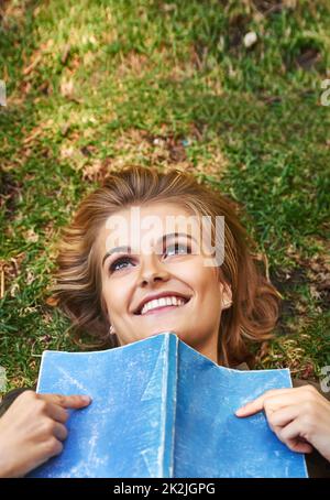 Books can really get your mind going. Shot of an attractive young woman reading a book while lying outside on the grass. Stock Photo