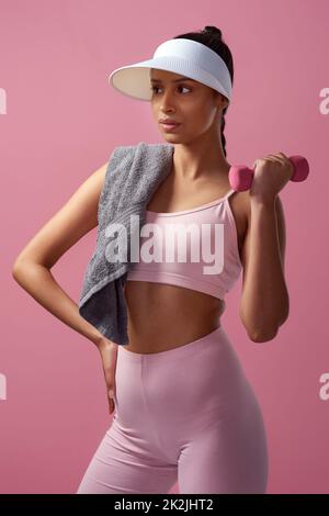 Fitness is a lifestyle. Cropped shot of an attractive and sporty young woman posing with a towel and a dumbbell in studio against a pink background. Stock Photo