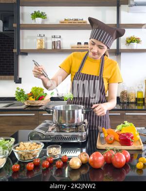 Young asian housewife dressed in an apron and a hair cap, preparing the vegetable soup in a modern kitchen. Stock Photo