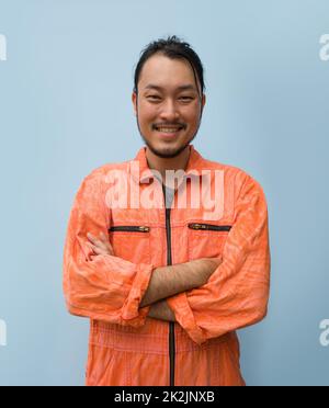 Young long hair asian man with mustache and beard stand smiling with his arms crossed. Portrait of industrial people on blue background. Stock Photo