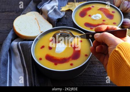 Female in yellow knitted sweater eating pumpkin cream soup on dark wooden background with spoon . Autumn cozy dinner concept Stock Photo