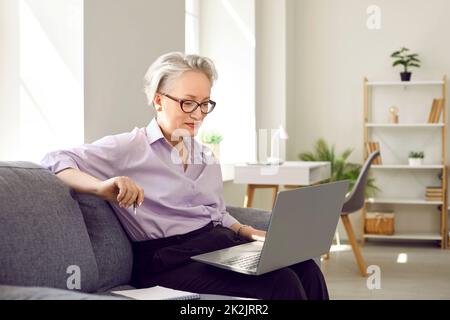 Senior woman sitting with laptop on couch at home and having video call or watching webinar Stock Photo