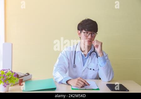 Young medical doctor in white coat thinking and looking seriously at hospital workplace. Stock Photo