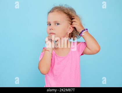 Portrait of cherubic blue-eyed little girl titivating touching short curly fair hair, wearing pink jumpsuit, bracelets. Stock Photo