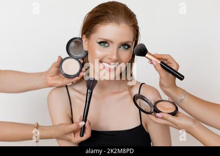 Photo closeup of pretty smiling girl with evening makeup and highlighting cheekbones on white background. Makeup artists applying makeup with brushes Stock Photo