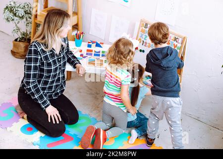 Little kids and educator studying numbers and alphabet on magnetic board sitting on floor in playroom backview. Interesting lesson for kindergartners Stock Photo