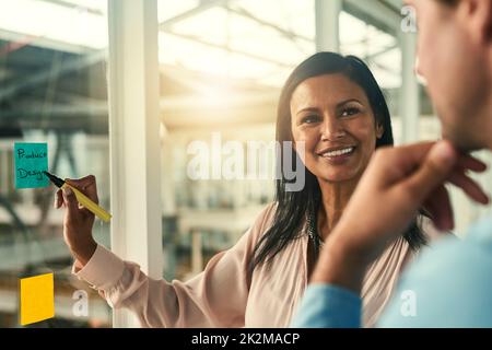 Bringing her vision to life. Shot of a confident businesswoman presenting an idea to her colleague using adhesive notes on a glass wall in the office. Stock Photo