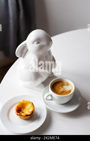 Easter Bunny stands near a cup of cappuccino and a cake on a plate, a white cup of coffee. Easter table decoration. copy space. Stock Photo
