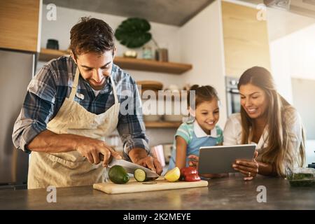 Cooking by their instructions. Shot of two happy parents and their young daughter trying a new recipe in the kitchen together. Stock Photo