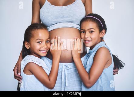 Lets have a listen. Portrait of two cheerful little girls standing next to their pregnant mother at home during the day. Stock Photo
