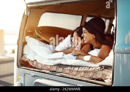 Anywhere is better together. Shot of two happy friends relaxing on a blanket in the true of their van on a road trip. Stock Photo