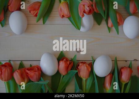 Pink tulips with green leaves lie in a row at the bottom and top on a wooden background with a place for text, white Easter eggs are cut next to them Stock Photo