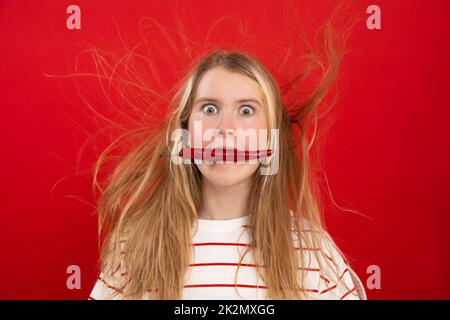 Portrait of shocked teenage girl with flying disheveled fair hair, wearing white T-shirt, keeping in mouth red chilli. Stock Photo