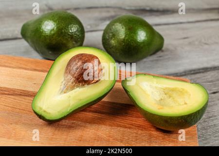 Avocado cut in half, seed visible, on wooden chopping board, two more whole pears in background. Stock Photo