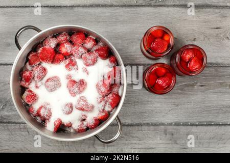 Tabletop view - large steel pot with strawberries covered in crystal sugar, three pickled strawberry glass bottles next to it. Homemade compote preparation. Stock Photo