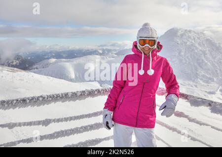 Young woman in pink jacket, wearing ski goggles, leaning on snow covered fence, smiling, white mountains in background. Stock Photo