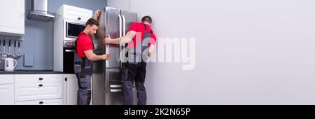 Two Young Male Movers Placing Steel Refrigerator In Kitchen Stock Photo