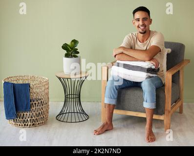 I love folding laundry. Shot of a young man sitting in a chair folding laundry at home. Stock Photo