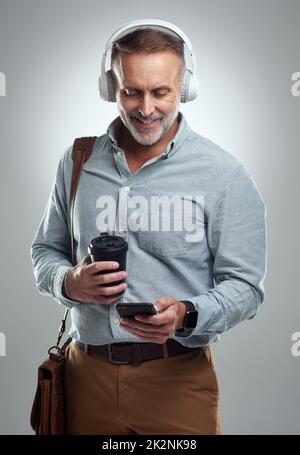 Hes a go-getter whos always prepared for the day. Studio shot of a mature man wearing headphones and using a cellphone while carrying a bag and cup of coffee against a grey background. Stock Photo