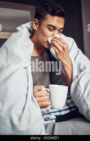 Dont get the flu, get the shot. Shot of a young man blowing his nose and having tea while recovering from an illness in bed at home. Stock Photo