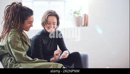 Two women friends using smartphone sitting on sofa at home browsing internet watching online entertainment on mobile phone Stock Photo