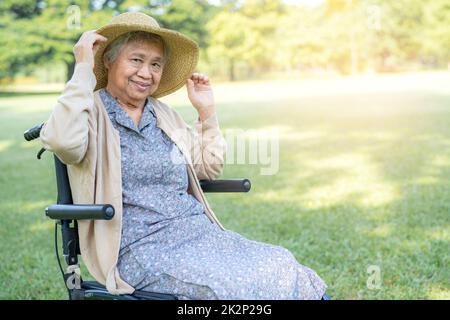 An elderly woman traveler with straw hat sitting on wheelchair in park. Stock Photo