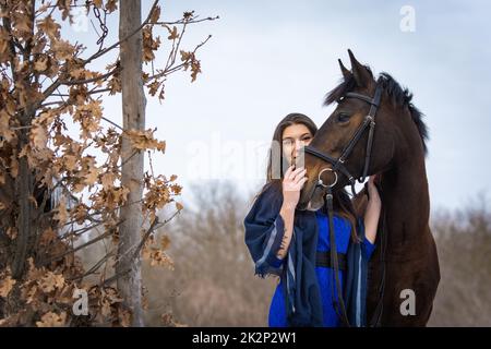 The girl peeks out from behind the muzzle of a horse, in the background an autumn forest Stock Photo