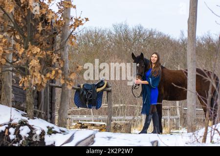 A girl in a blue dress walks with a horse through a farm in winter Stock Photo