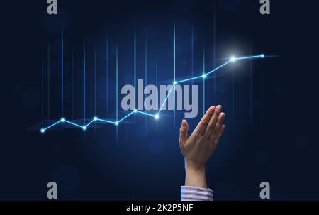 a businessman's hand points to a holographic graph with rising rates on a blue background. Development of a business with high profits, the prospect of implementing business plans Stock Photo