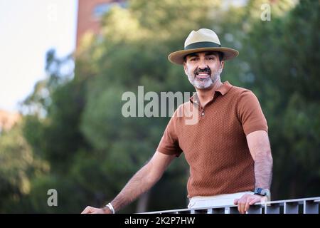 Middle-aged man in hat looking at camera and smiling while leaning on a railing outdoors. Stock Photo