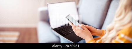 Woman with smartphone in her hands sitting on the couch and working on the laptop Stock Photo