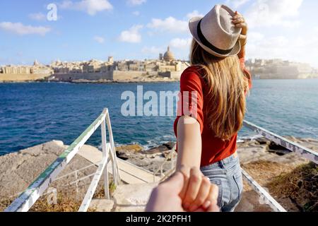 Follow me in Malta. Young carefree tourist woman in hat holds boyfriend hand and looking panoramic view of Valletta, Malta. Couple on summer holiday vacation. Traveling together. Stock Photo