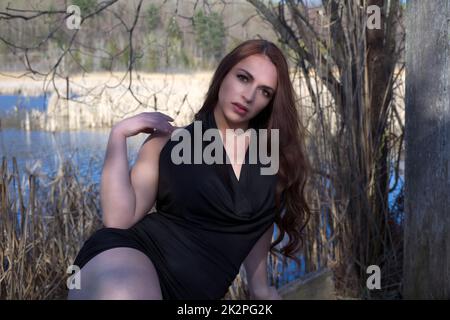 young woman in black dress sitting in the forest near water pond in sunshine Stock Photo
