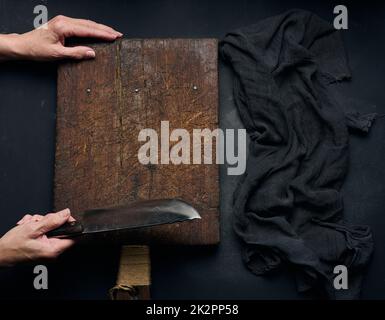 empty brown rectangular wooden cutting board, woman holding big knife over black table, top view Stock Photo