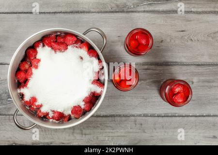 Tabletop view, large steel pot full of strawberries covered with crystal sugar, three bottles of strawberry in syrup next to it on gray wood desk. Homemade compote preparation. Stock Photo