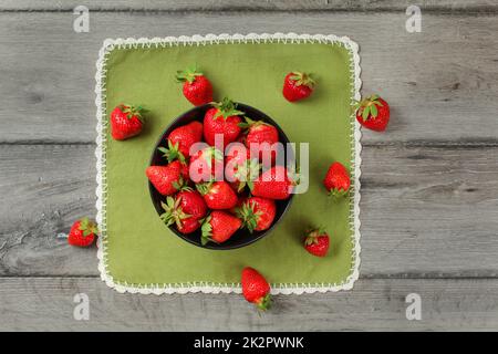 Tabletop view, small black ceramic bowl, with strawberries, some of them spilled on green tablecloth under. Stock Photo