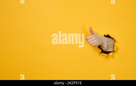 woman's hand sticks out of the torn hole and shows a gesture okay, thumb up. Stock Photo