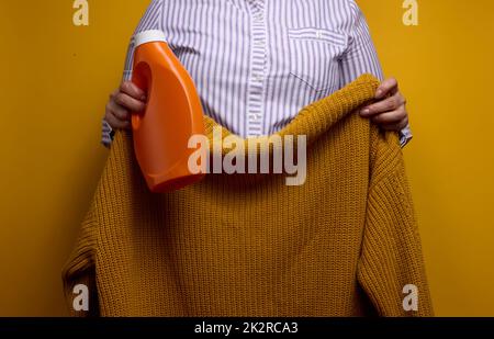a woman in a white striped shirt holds an orange plastic bottle with liquid washing gel and a knitted sweater. Laundry and household chores, clean washed items Stock Photo