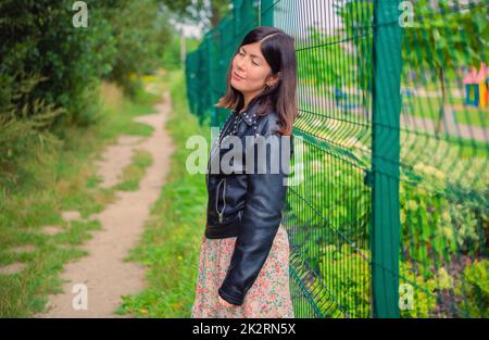 A cute girl in a black jacket stands with her eyes closed, leaning her back against a green iron fence. Stock Photo